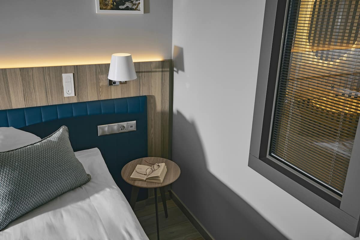 Ein Hotelzimmer mit Bett, Nachttisch mit Lampe, Fenster mit Jalousien und einem fest montierten Lichtschalter.