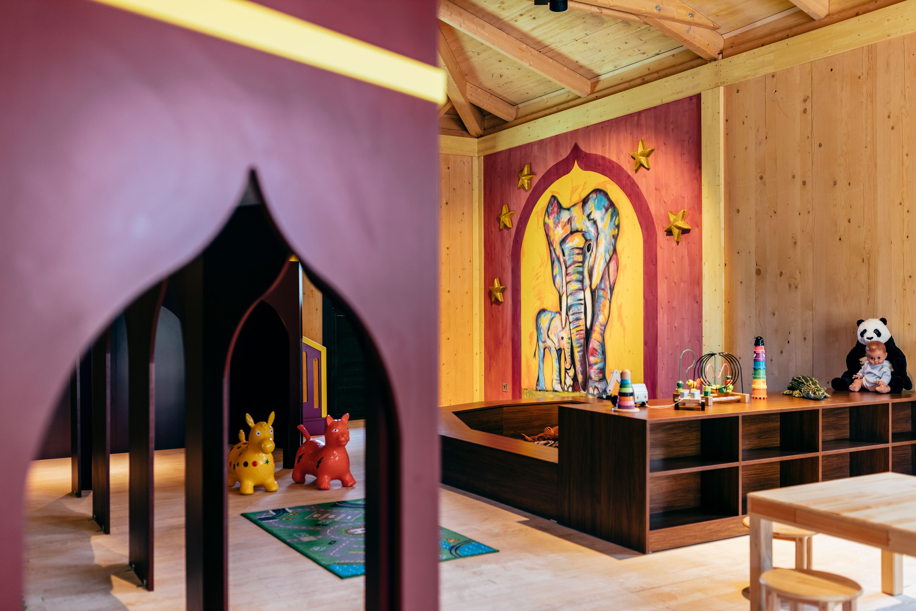 A room with a wooden ceiling, a colorful mural, a play area with toys, and a table.