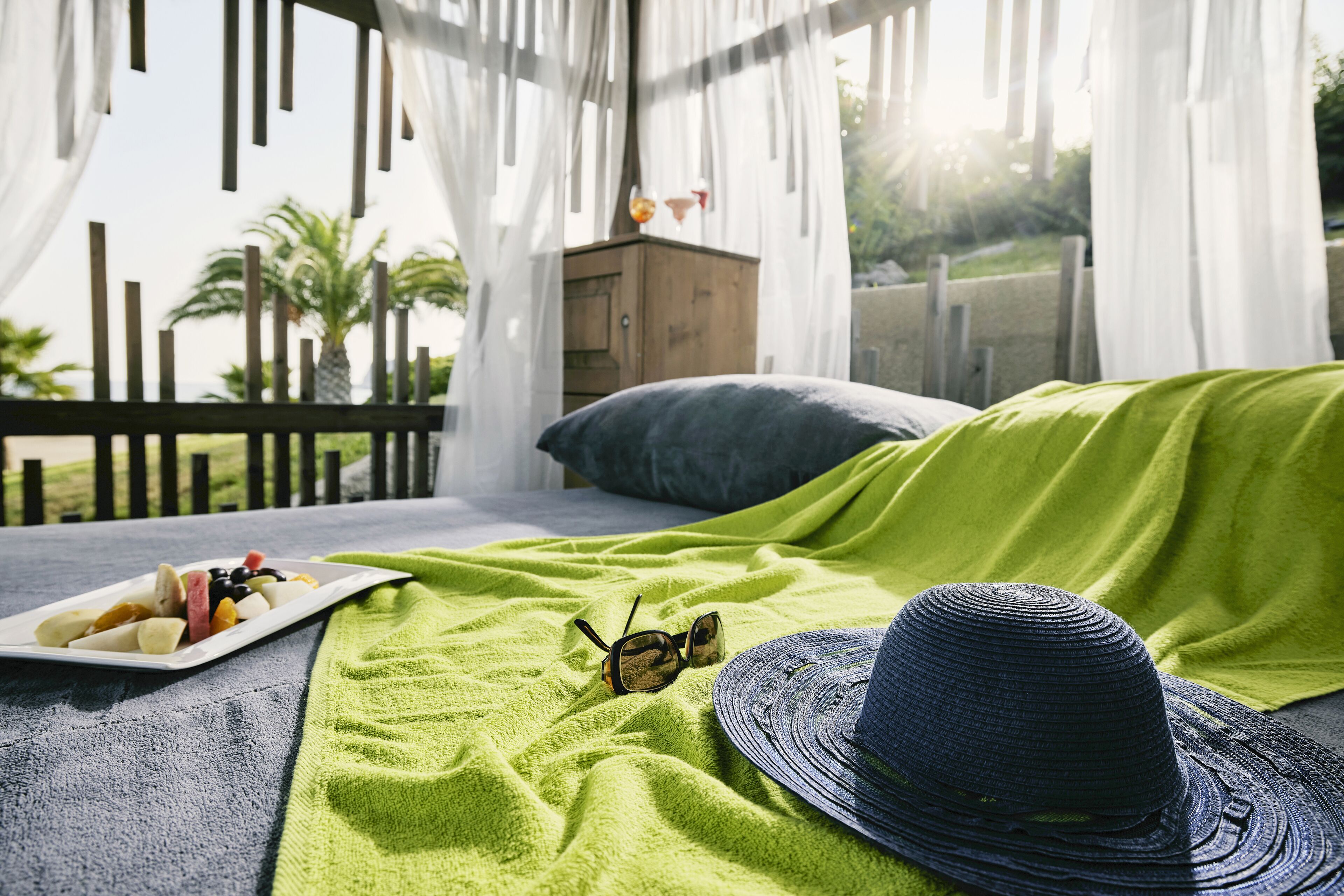 Una cama con una manta verde, un plato de fruta, gafas de sol y un sombrero en un balcón con baranda de madera y vistas a palmeras.