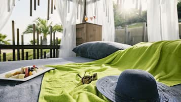 Una cama con una manta verde, un plato de fruta, gafas de sol y un sombrero en un balcón con baranda de madera y vistas a palmeras.