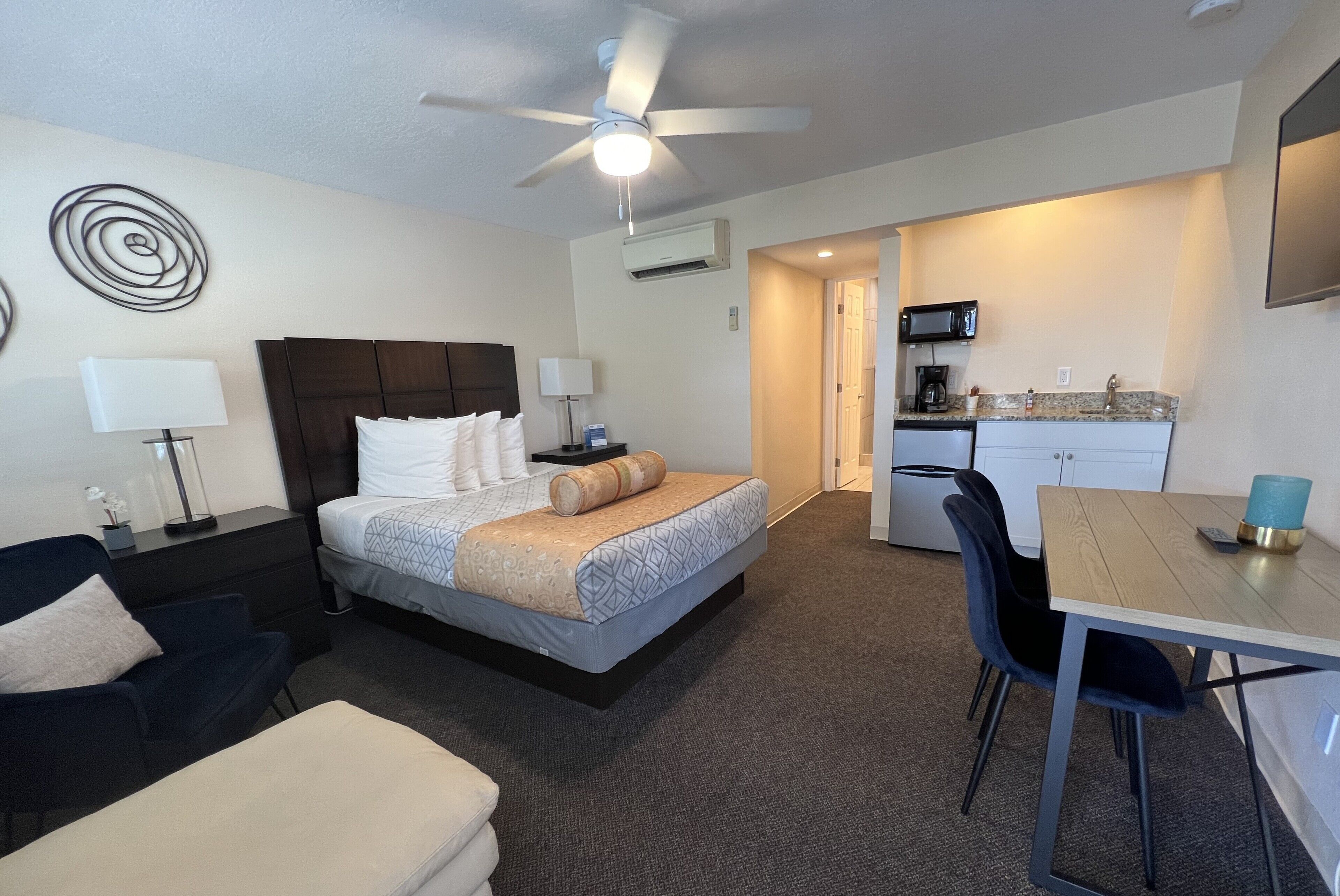 A hotel room with a bed, a desk, a chair, a ceiling fan, and a kitchenette.