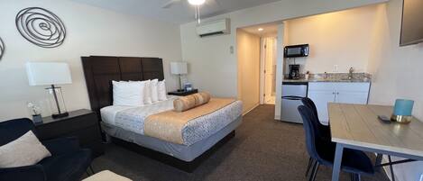 A hotel room with a bed, a desk, a chair, a ceiling fan, and a kitchenette.