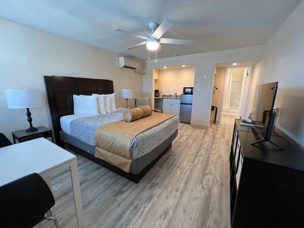 Habitación de hotel con cama, mesitas de noche, escritorio, silla, ventilador de techo, equipo de aire acondicionado, kitchenette y espejo.