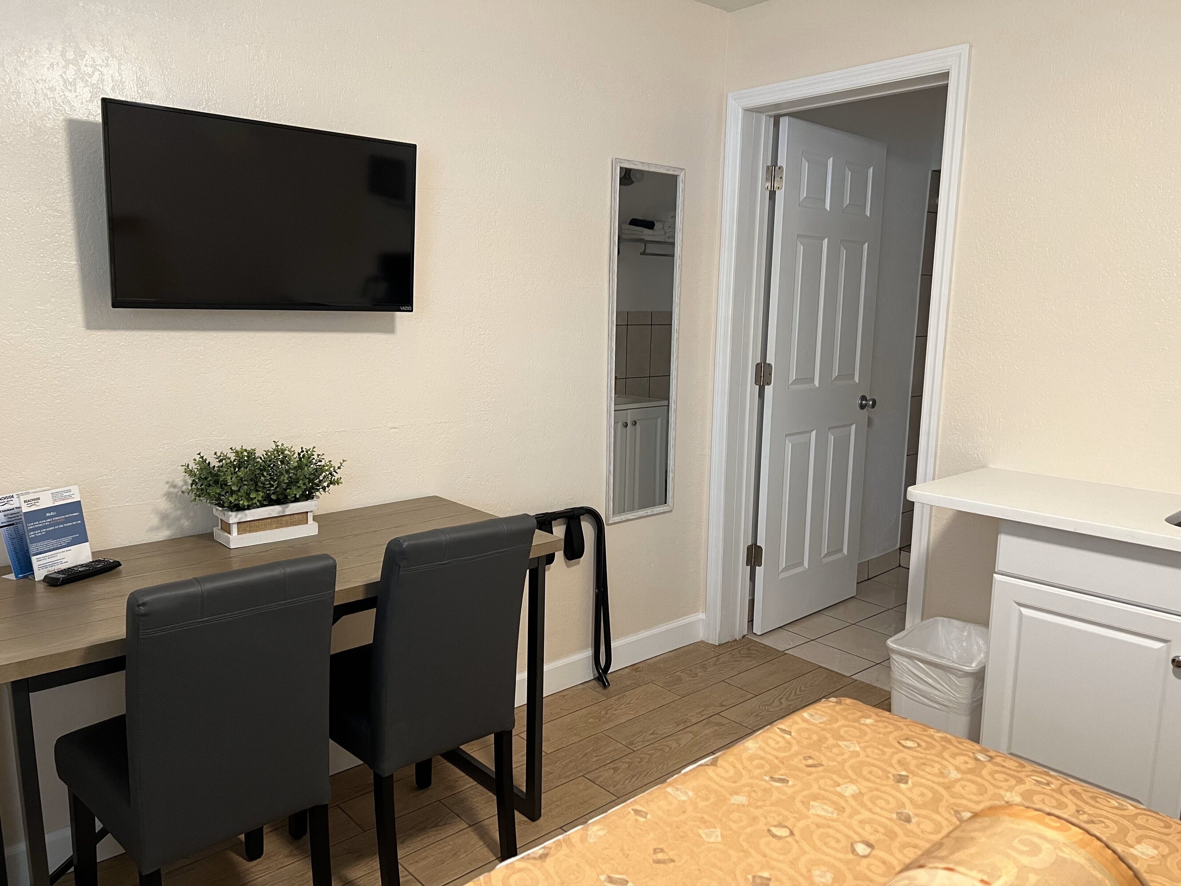 A room with a wooden table, two black chairs, a small potted plant, a flat-screen TV mounted on the wall, a white door, and a white cabinet.