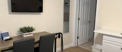 A room with a wooden table, two black chairs, a small potted plant, a flat-screen TV mounted on the wall, a white door, and a white cabinet.