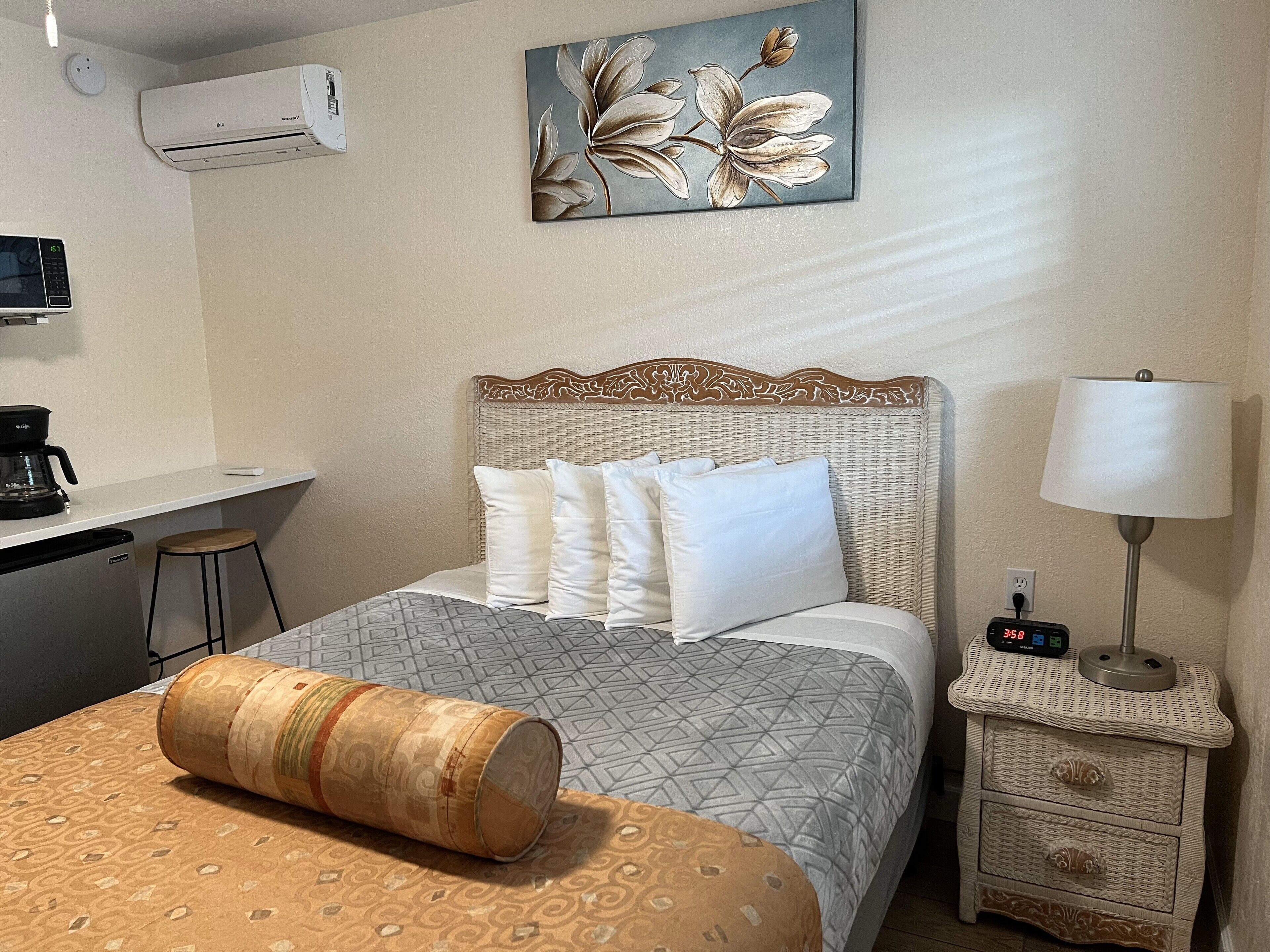 A hotel room with a bed, a bedside table with a lamp, a small desk with a coffee maker, and an air conditioning unit.