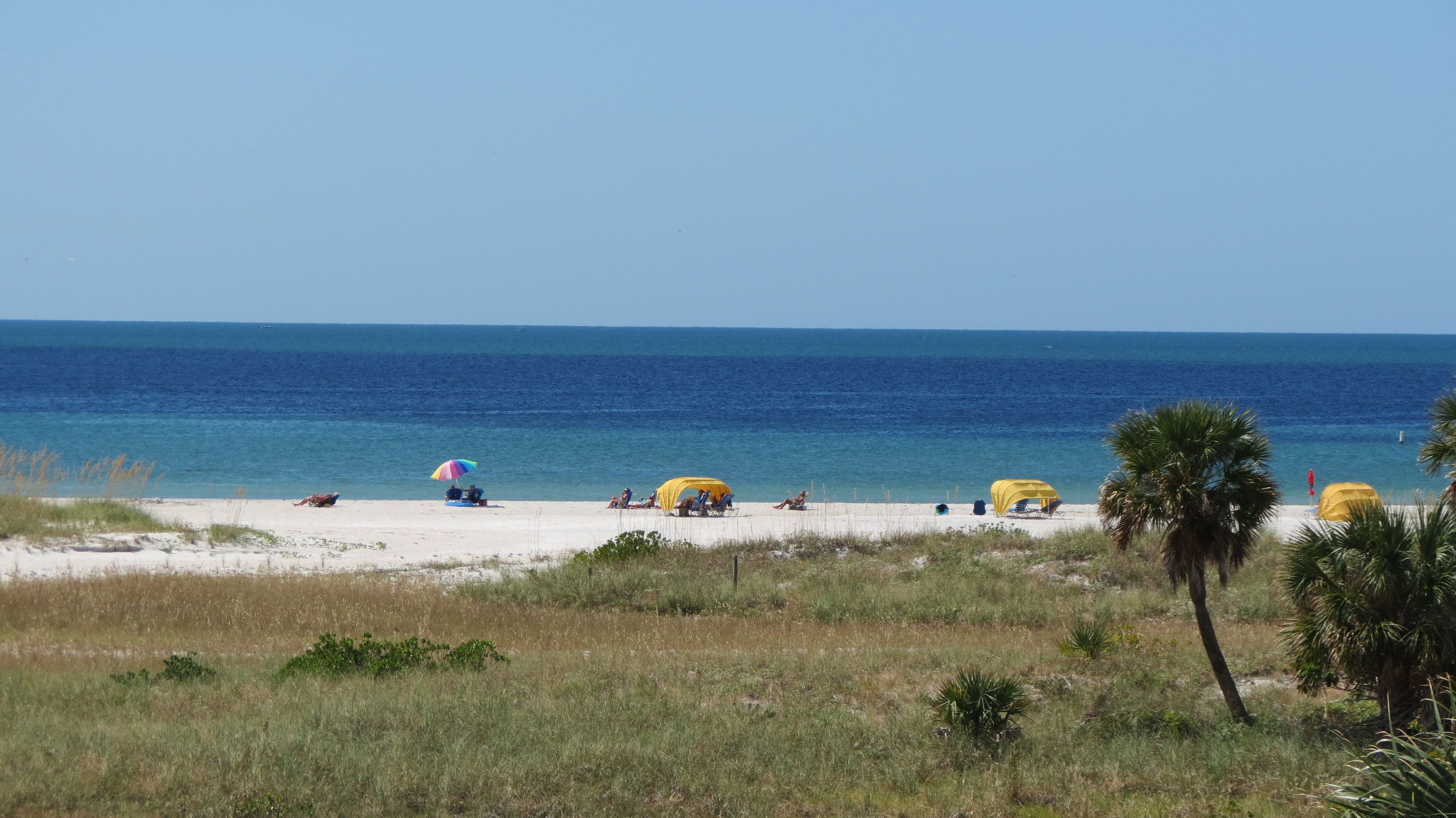 Een strand met wit zand, helderblauw water en enkele gele strandparasols.