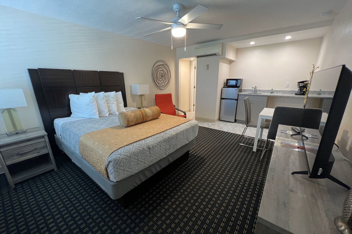 A hotel room with a bed, a desk, a chair, a ceiling fan, a TV, and a kitchenette.