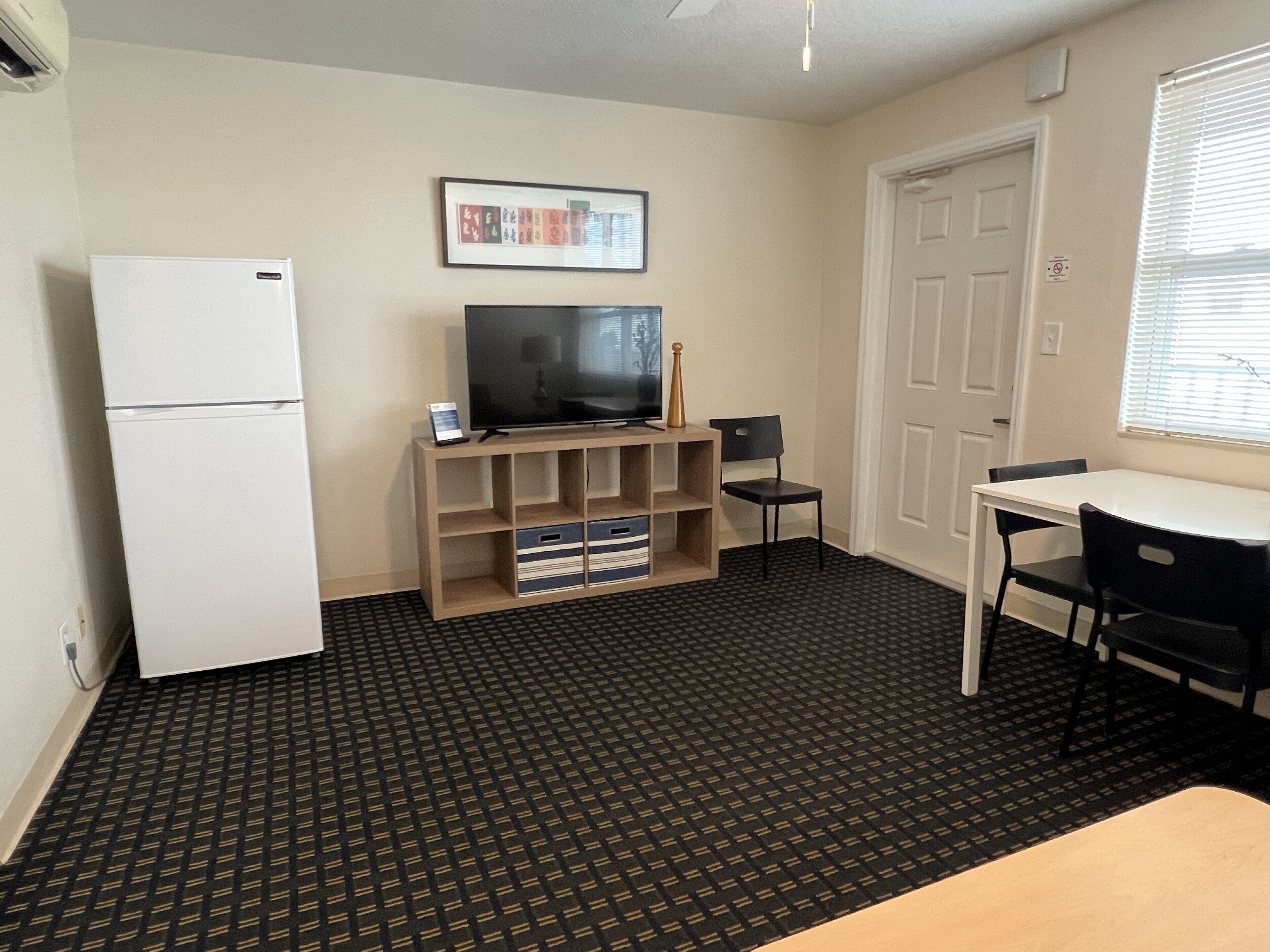 A compact room with a refrigerator, a TV on a wooden stand, a dining table with chairs, and a patterned carpet.