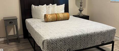 A bedroom with a bed, a nightstand, a lamp, a framed picture, and a window with blinds.