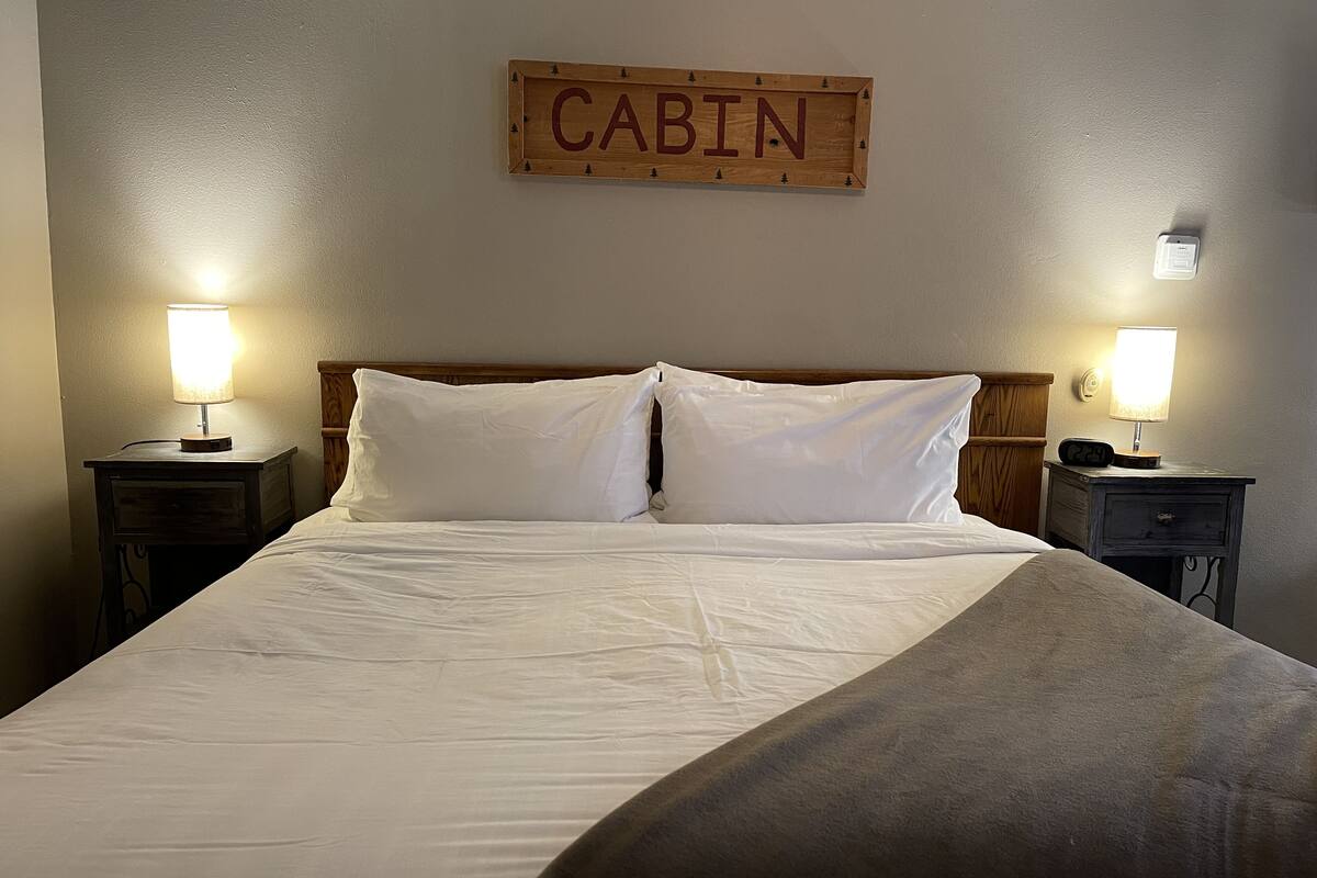 A neatly made bed with white linens, two bedside lamps, and a wooden sign above reading "CABIN".