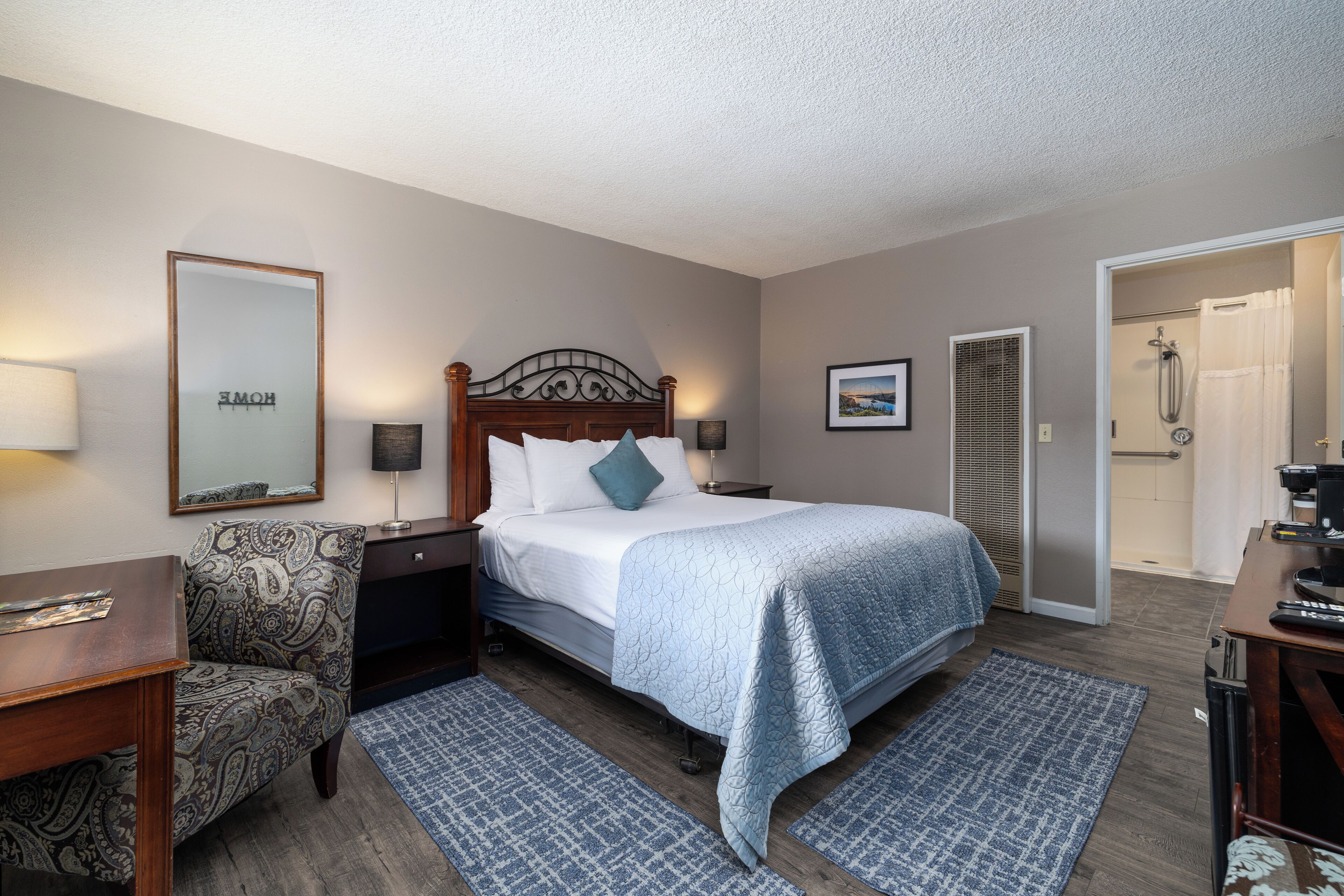 A hotel room with a bed, a chair, a desk, a mirror, and a shower area.