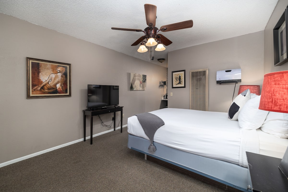 A hotel room with a bed, ceiling fan, artwork on the walls, a television, and an air conditioning unit.