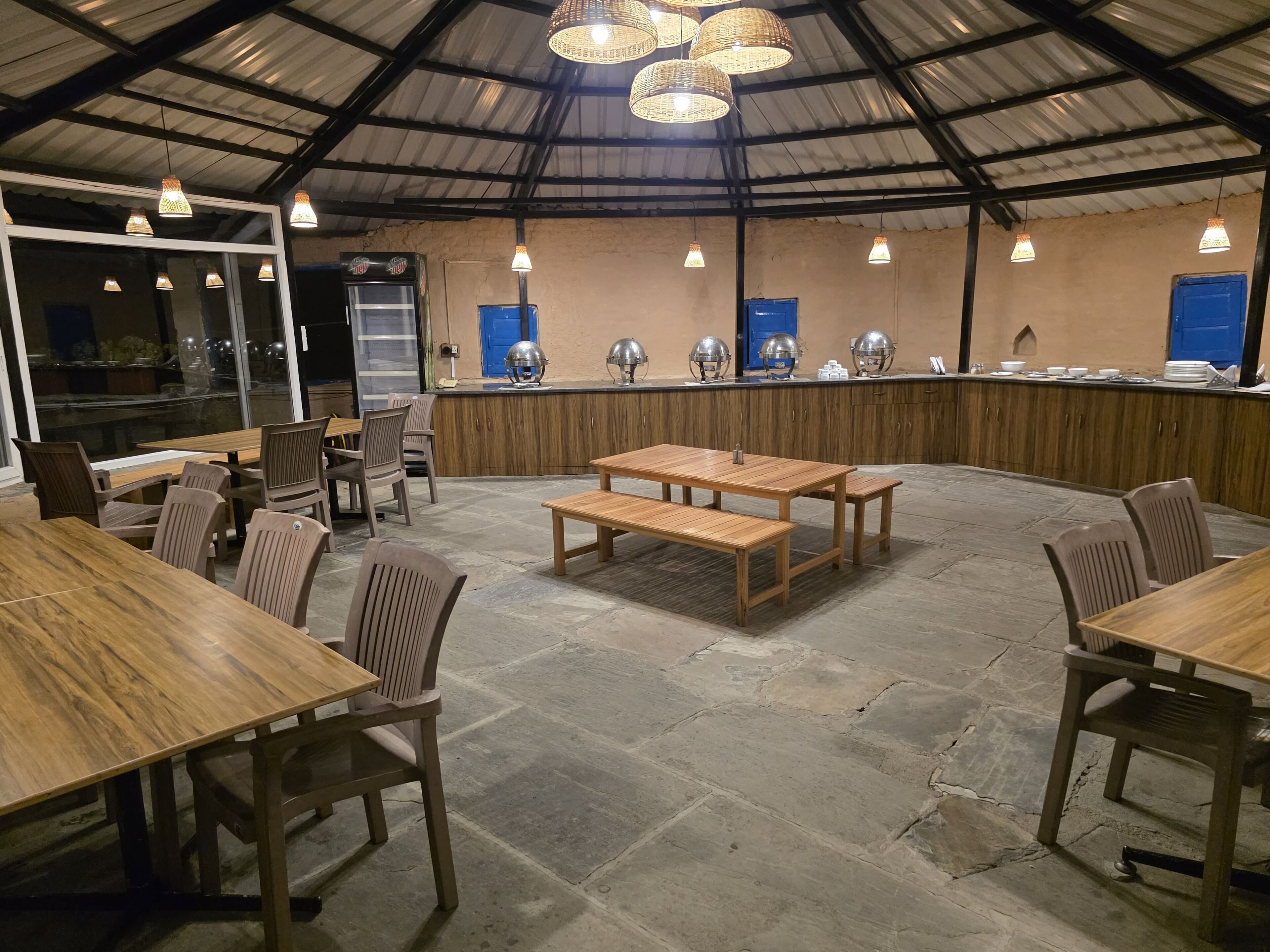 An indoor dining area with wooden tables and chairs, a counter with food items, and a view through large windows.