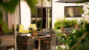 An outdoor seating area with wicker furniture and a table, surrounded by greenery and a building with large windows. - Adina Apartment Hotel Hamburg Michel (Hamburg)