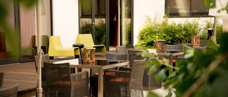 An outdoor seating area with wicker furniture and a table, surrounded by greenery and a building with large windows.