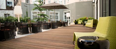 Una terraza de madera con sillas de mimbre y una mesa, rodeada de follaje y un paraguas grande.