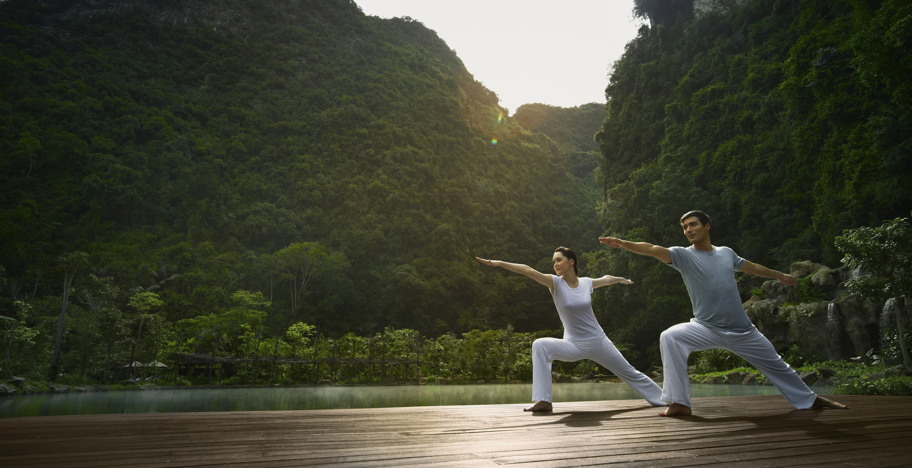 Two people practicing yoga on a wooden deck with a forest and waterfall in the background.