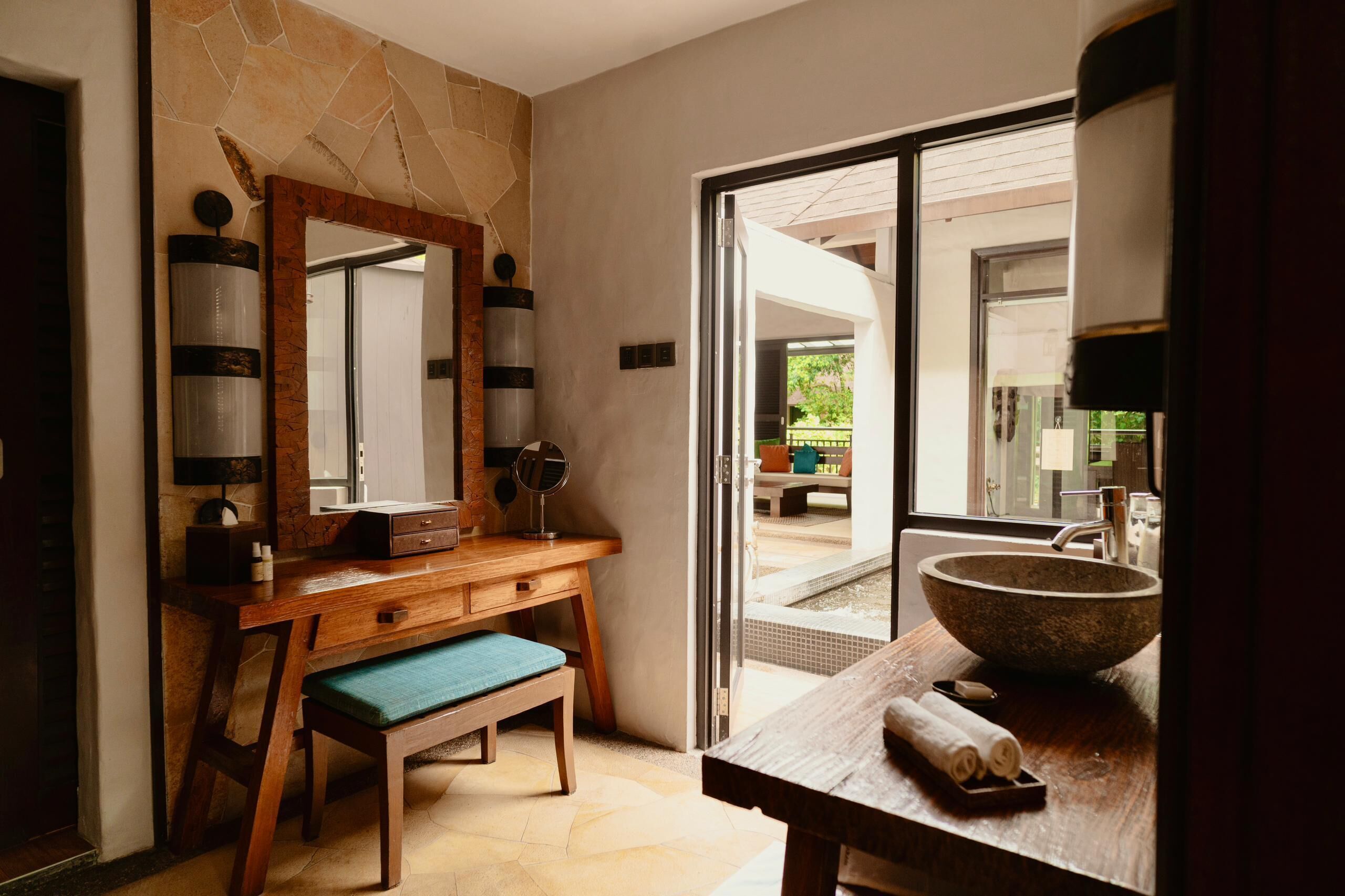 A bathroom with a wooden vanity, a large mirror, a stone sink, and a bench.