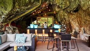 An outdoor bar area with a rustic stone wall backdrop, wicker furniture, and a bar counter with a bartender. - The Banjaran Hotsprings Retreat (Ipoh)