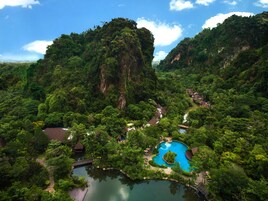 A lush, mountainous landscape with a clear blue pool surrounded by dense greenery and a few buildings.