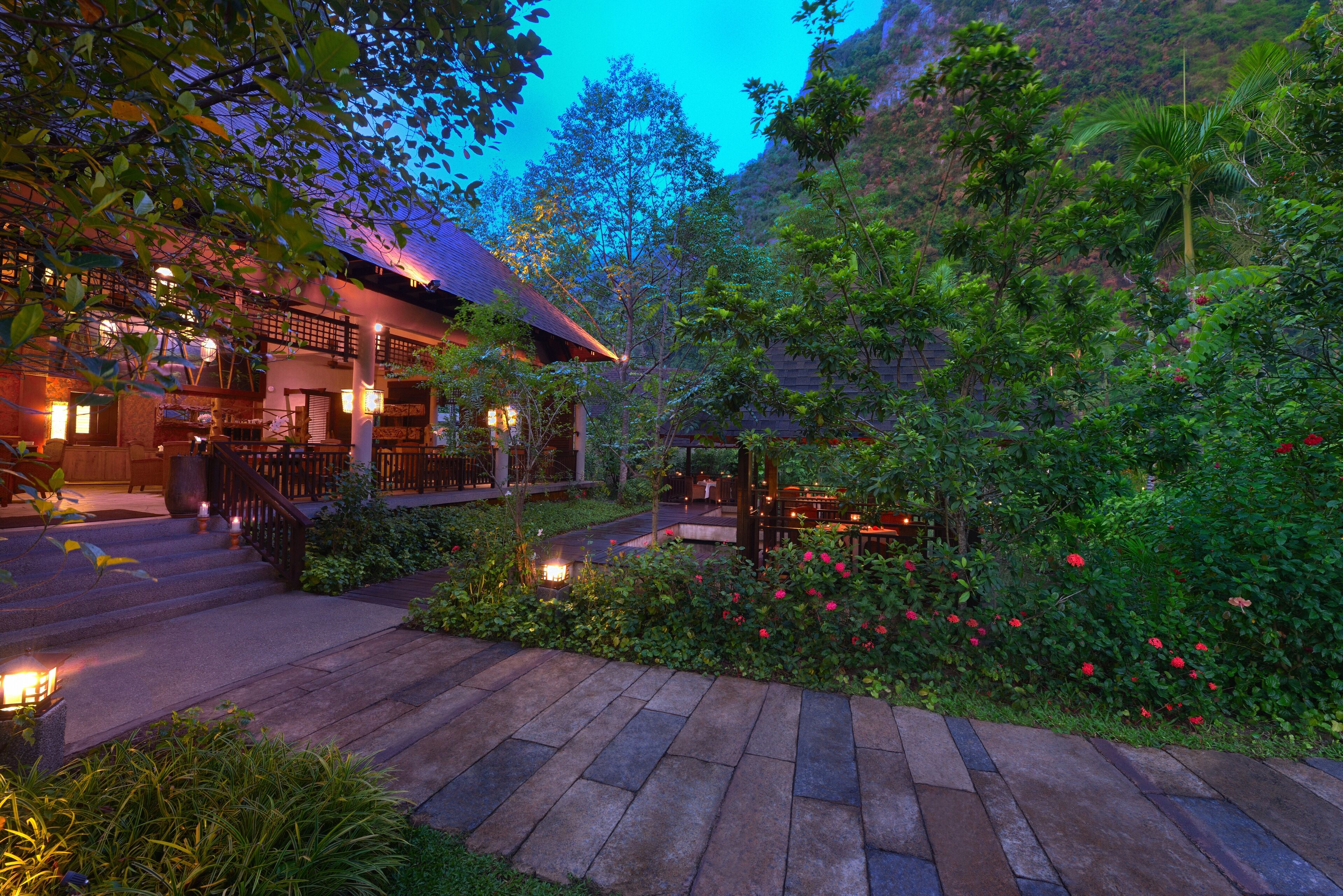 A traditional building with a wooden staircase leading to a porch, surrounded by lush greenery and flowering plants.