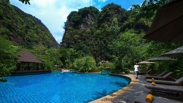 A swimming pool with a tiled edge, surrounded by lounge chairs and umbrellas, set against a backdrop of lush greenery and a mountainous landscape. - The Banjaran Hotsprings Retreat (Ipoh)