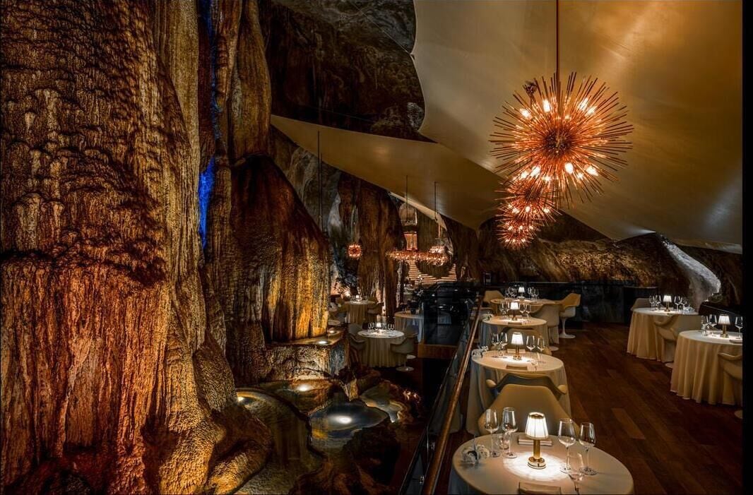 An interior with tables set for dining, a chandelier, and a large rock formation.