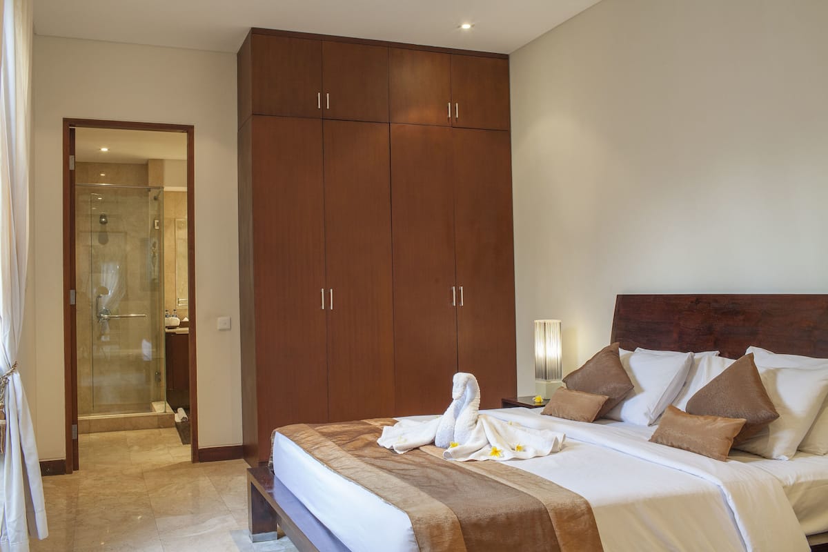 A hotel room with a large bed, a wooden wardrobe, a glass shower enclosure, and a bedside table with a lamp.