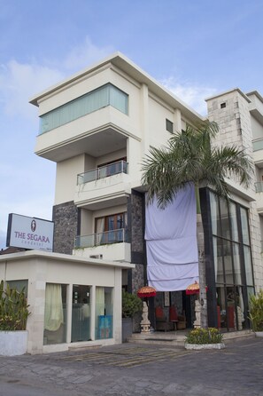 A multi-story hotel with a sign reading "The Segara Condomotel." It features a balcony with a palm tree, a covered entrance with red umbrellas, and a stone facade. - The Segara Suites (Nusa Dua)