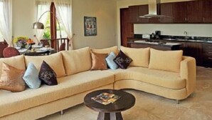 A modern living room with a beige sectional sofa, a round coffee table, and a kitchen area visible through an open doorway.