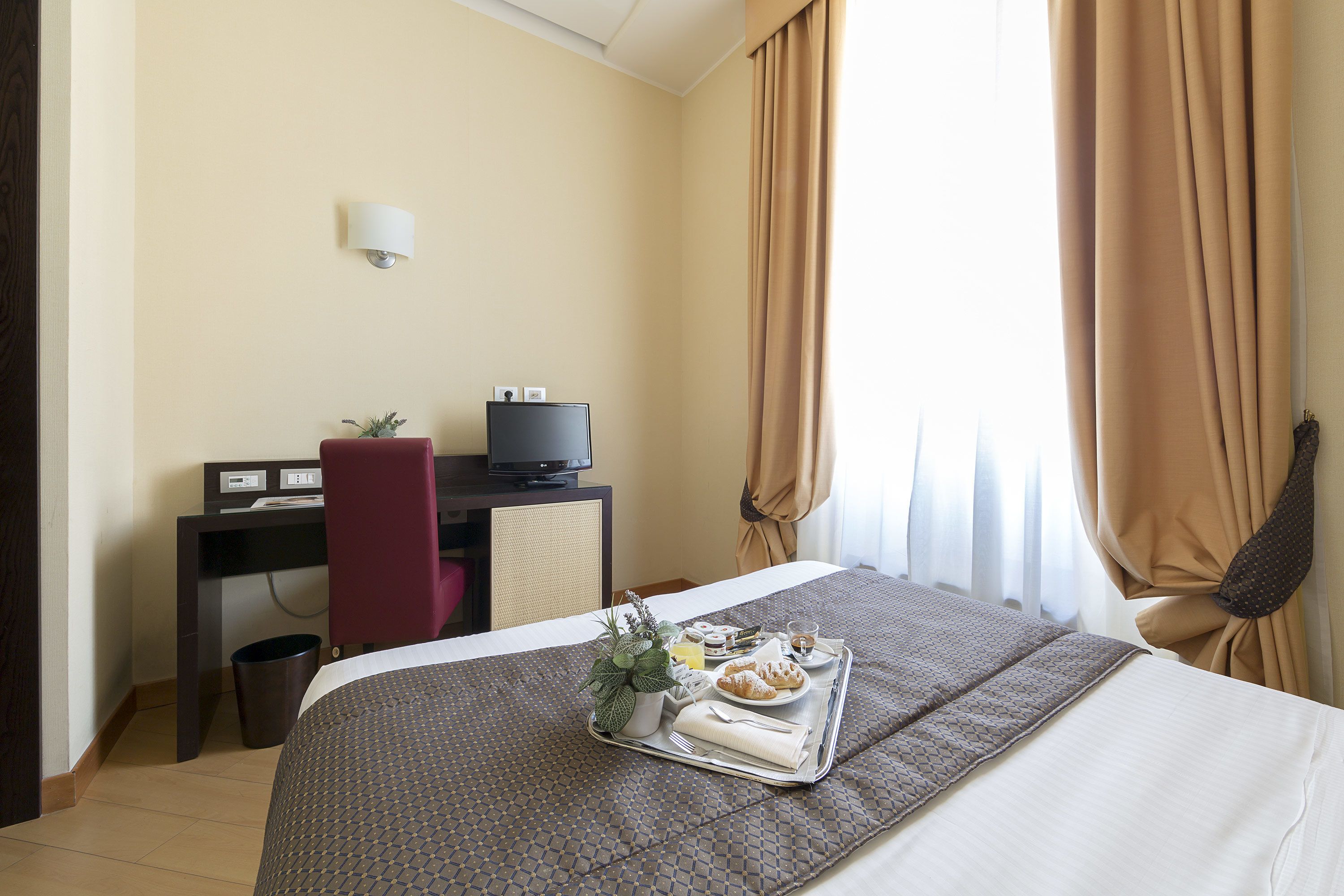 Camera d'albergo con un letto, una scrivania con un computer, un cestino e un vassoio con articoli per la colazione.