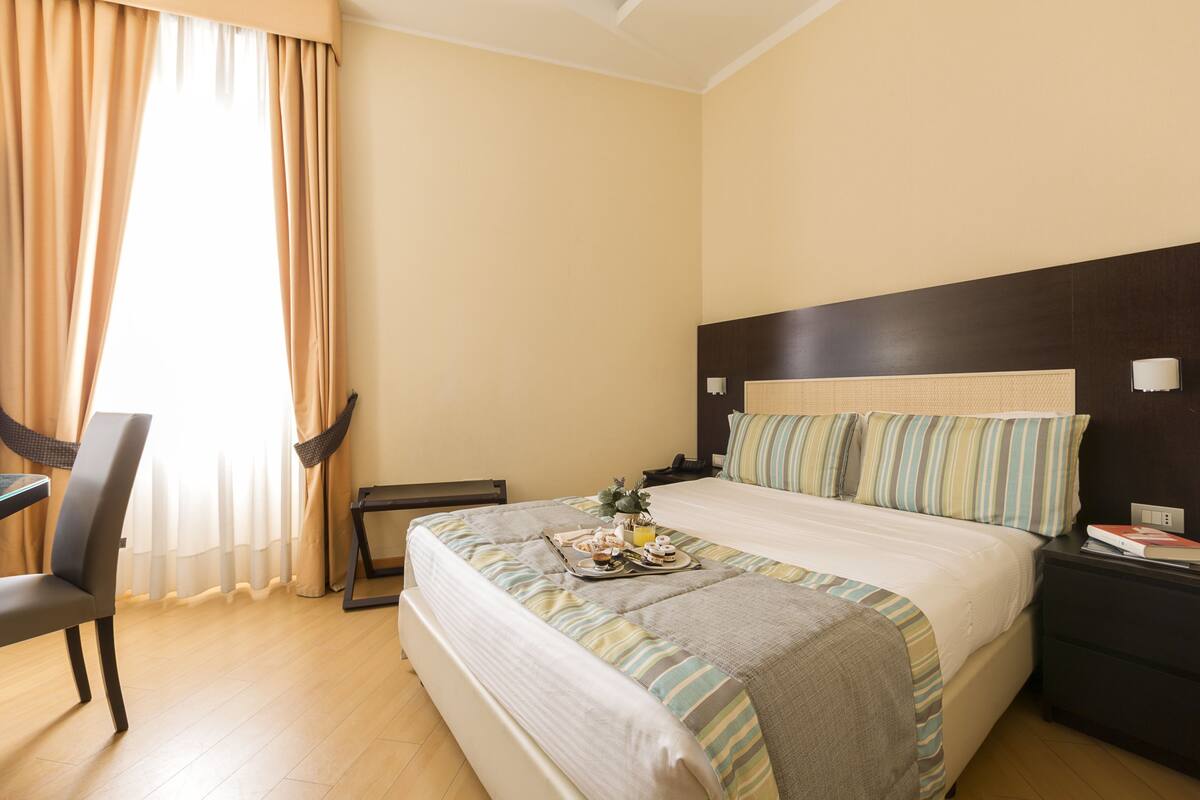 A hotel room with a bed, bedside table, chair, and a breakfast tray on the bed.
