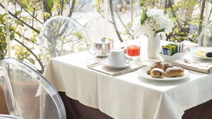 Una mesa de desayuno con mantel blanco, una jarra, una taza, un vaso de jugo y un plato con pasteles, además de un florero con flores.