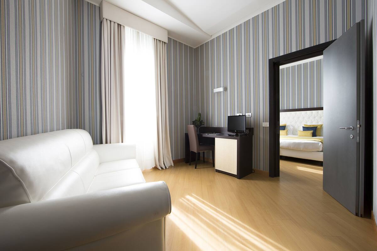 A modern hotel room with a white sofa, a desk with a computer, and a bed with yellow pillows.