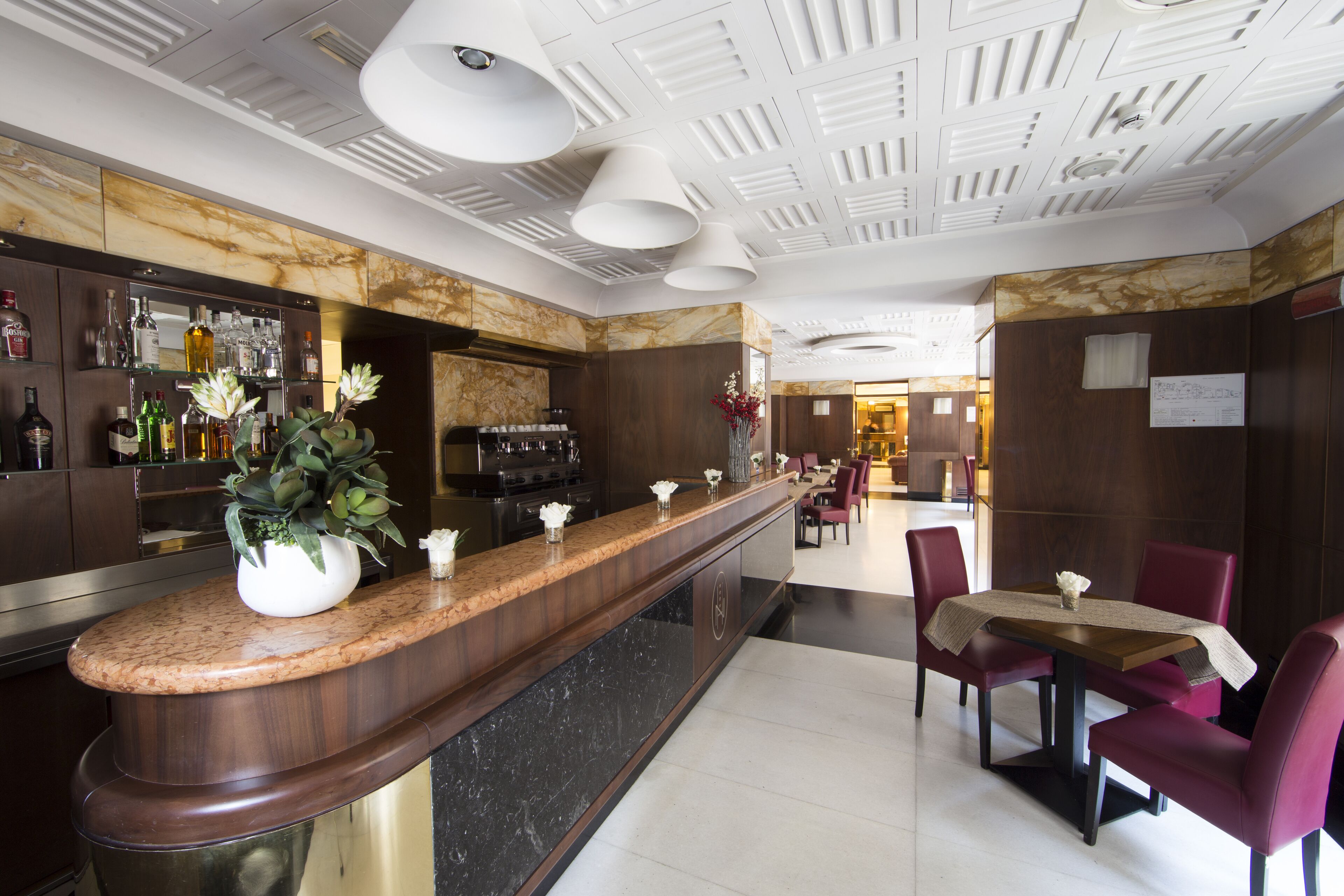 Um hall de hotel moderno com uma recepção de mármore, área de bar e um espaço de refeições com cadeiras vermelhas e uma mesa.