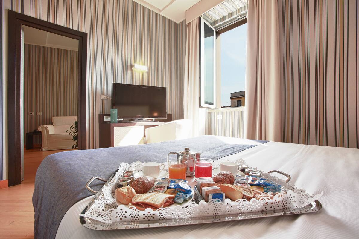 A hotel room with a bed, a TV, a breakfast tray with pastries and drinks, and a view of the outside through a window.