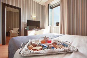 Une chambre d’hôtel avec un lit, une télévision, un plateau de petit-déjeuner avec des viennoiseries et des boissons, et une vue sur l’extérieur par une fenêtre.