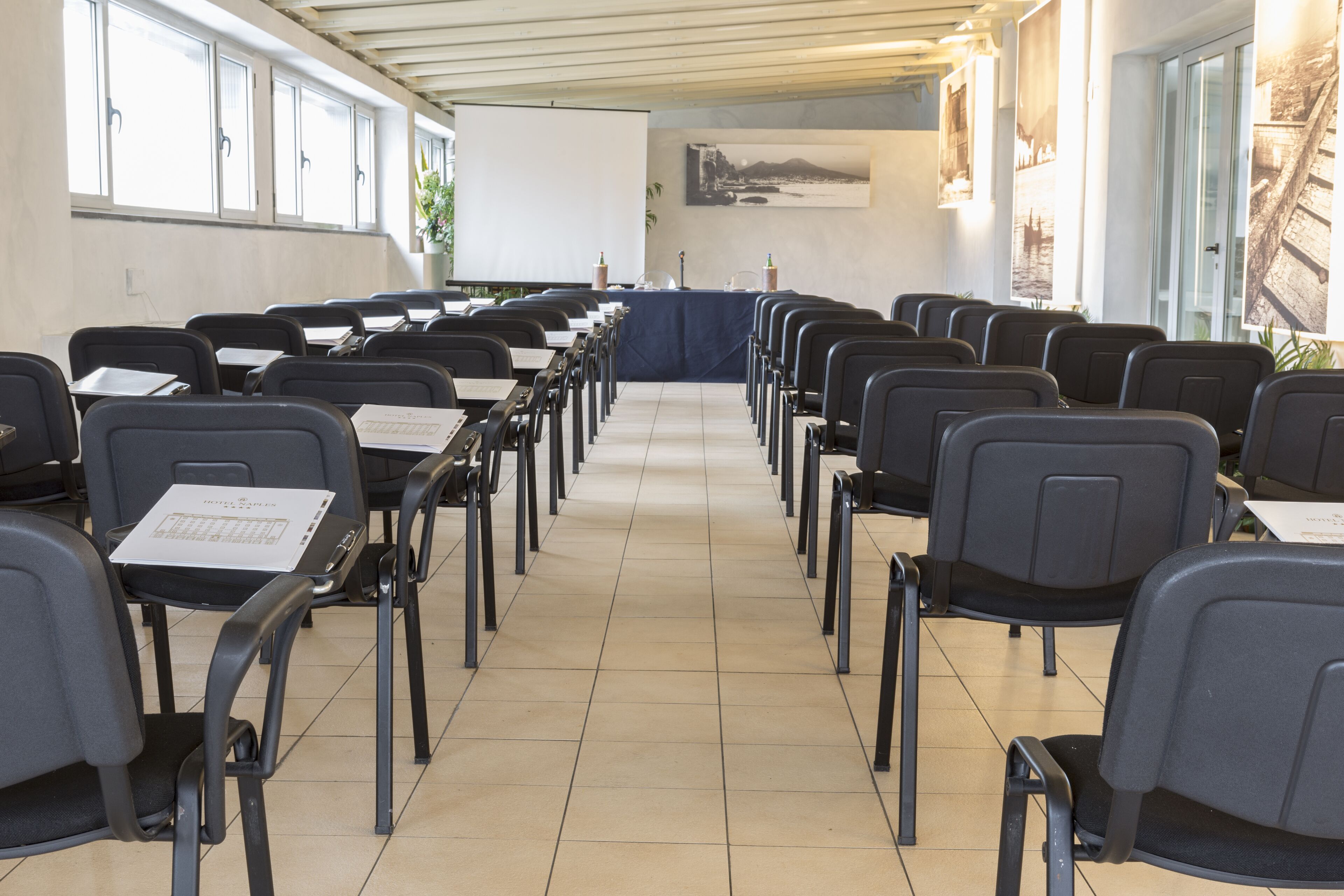 Een vergaderruimte met rijen stoelen en tafels, een projectiescherm en een groot raam met jaloezieën.