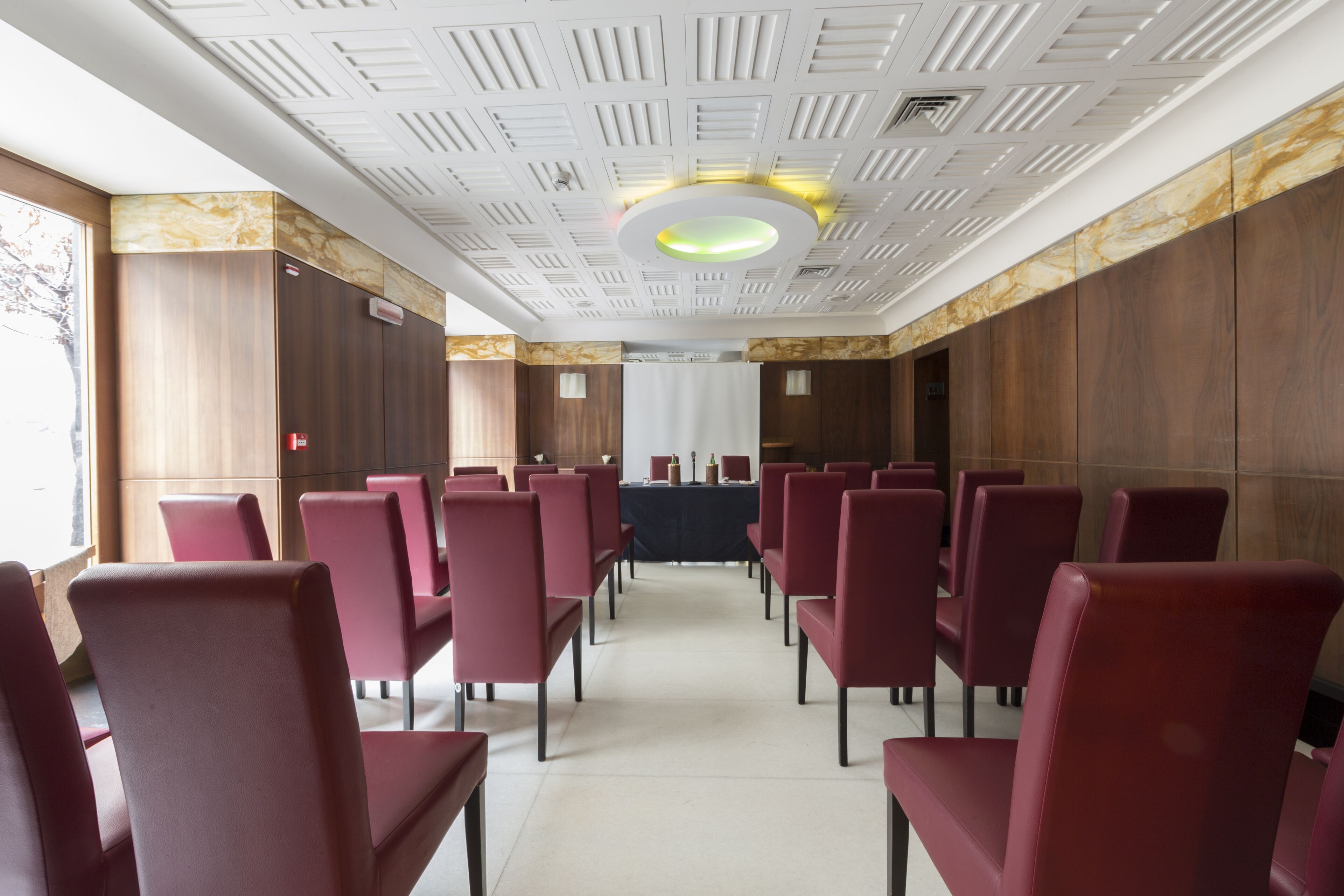 Une salle de conférence avec des rangées de chaises rouges, une table centrale et un plafond doté d’un éclairage encastré et d’un luminaire circulaire.