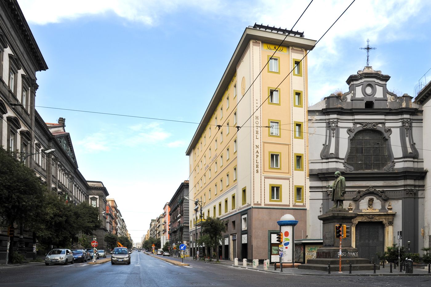 Straßenansicht mit einem Hotel namens 'HOTEL NAPLES' auf der rechten Seite, das eine klassische Fassade und eine Statue zeigt.