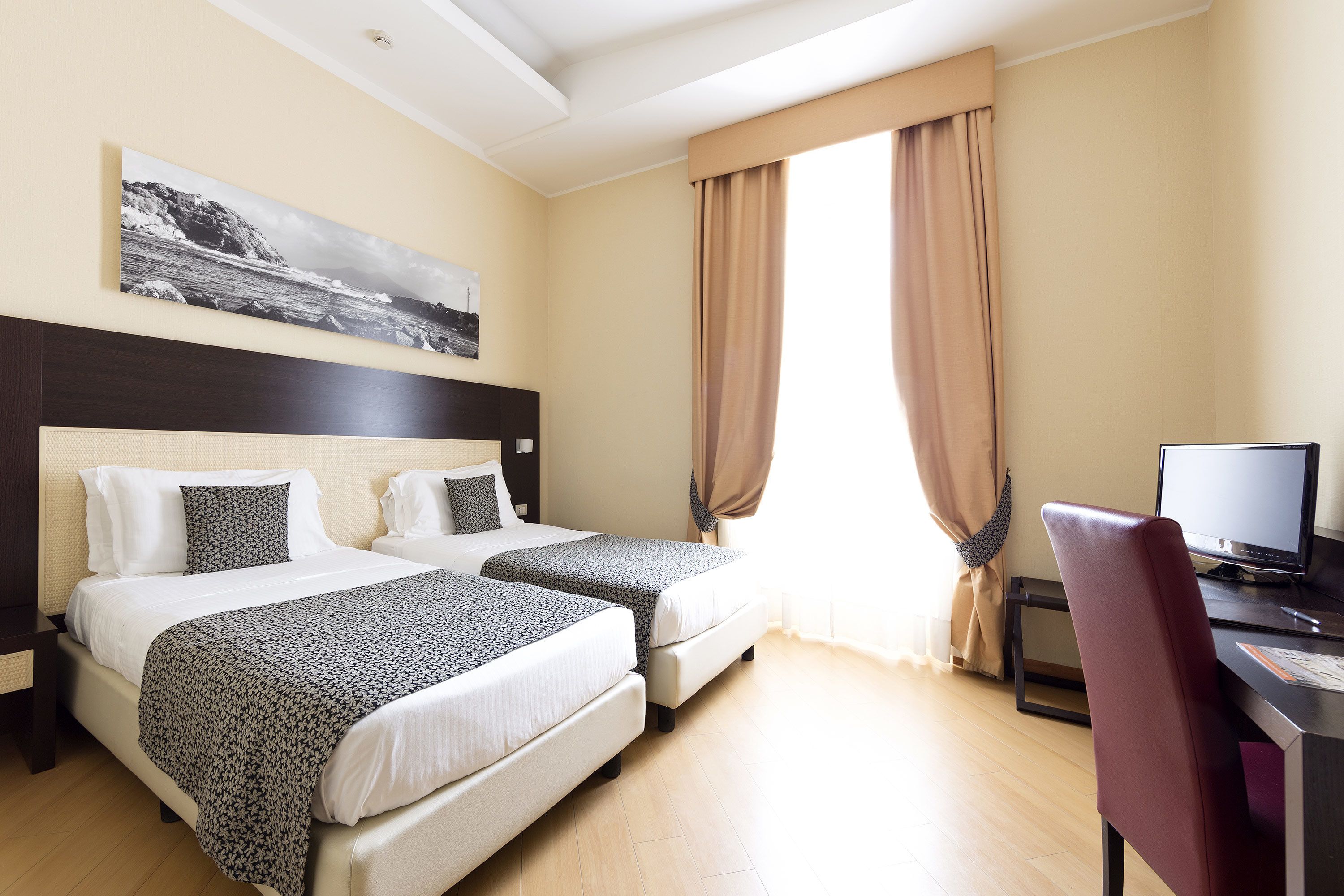 Une chambre d’hôtel avec deux lits, un bureau, un ordinateur et une grande fenêtre avec des rideaux.