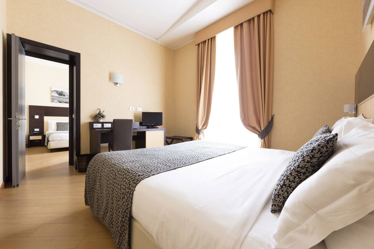 A hotel room with a bed, a desk, a chair, a TV, and a window with curtains.
