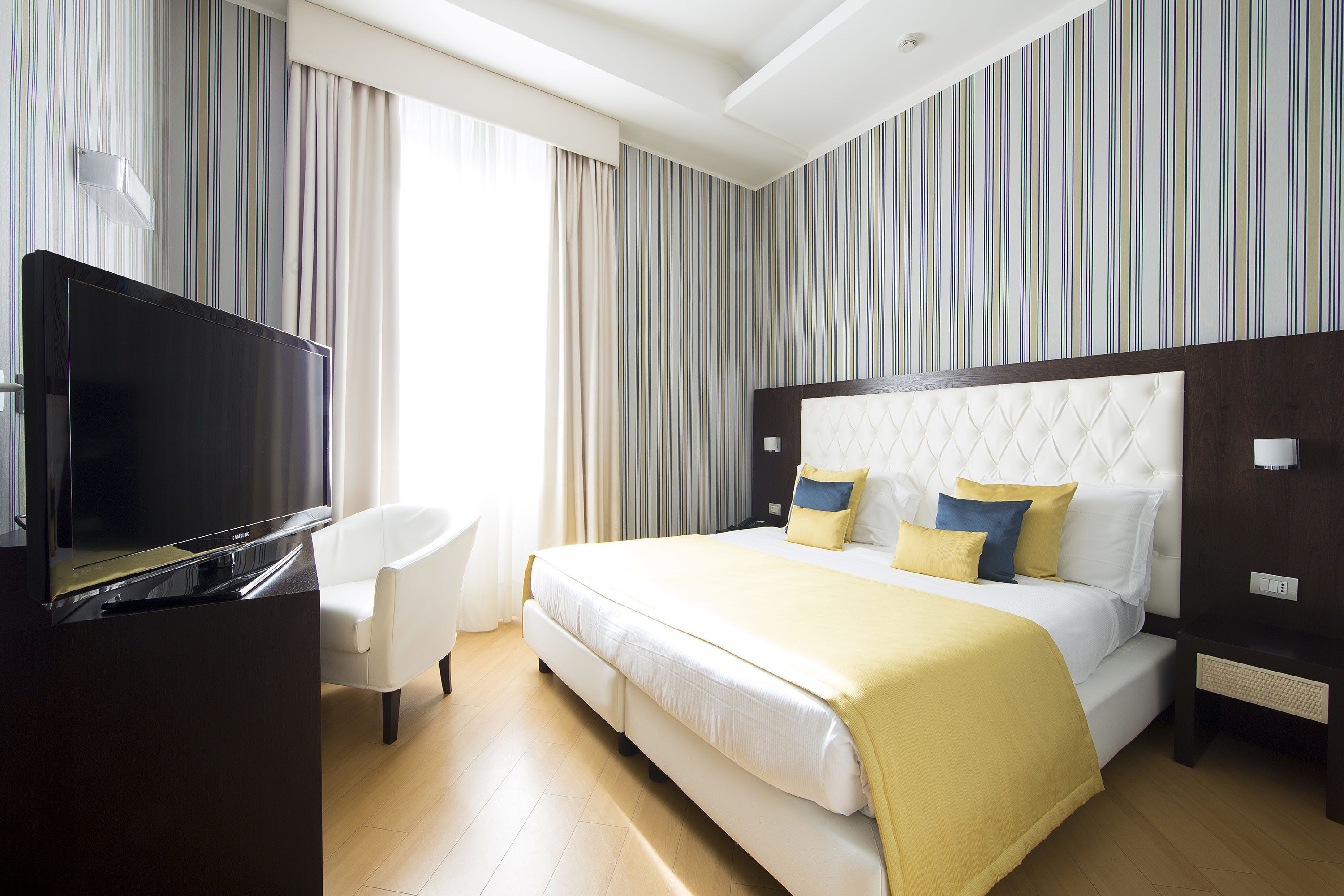 Una habitación de hotel con una cama grande, un televisor, un sillón blanco y papel tapiz a rayas.