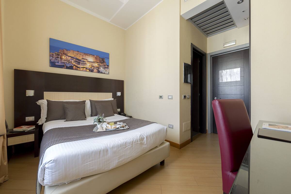 A hotel room with a large bed, a desk with a chair, a painting on the wall, and a door.