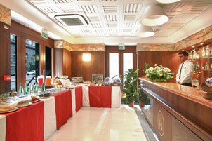 Un vestíbulo de hotel con mostrador de recepción, zona de buffet con diversas opciones de comida y un amplio ventanal con vistas a la calle.