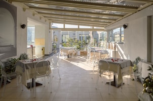 Un vaste espace de restauration intérieur, doté de tables et de chaises en verre transparent, d’un sol carrelé et de grandes fenêtres laissant entrer la lumière naturelle.