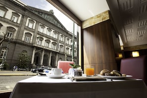 Una mesa de comedor con alimentos para el desayuno, vista por la ventana a un edificio histórico y una calle de la ciudad.