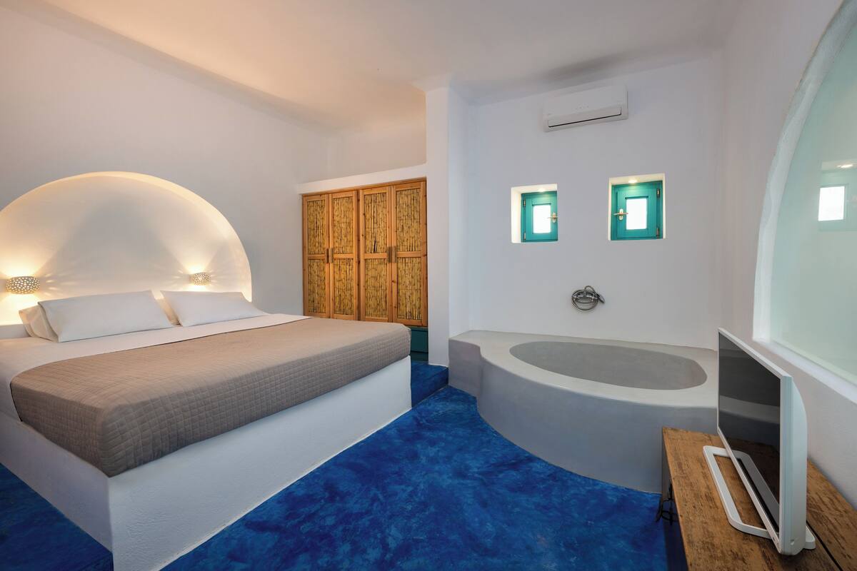 A bedroom with a bed, a large window, a wooden wardrobe, a bathtub, and a TV.