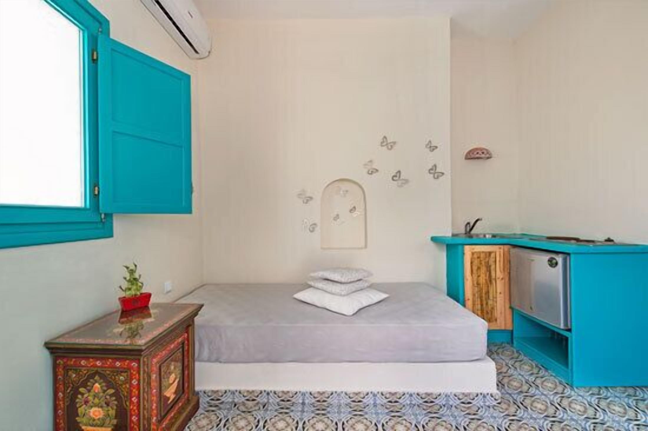 A small, cozy room with a bed, a small table with a plant, a wall-mounted shelf, and a window with blue shutters.